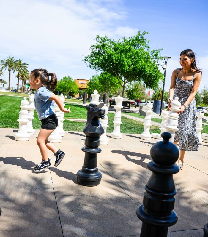 A woman and child play with giant chess pieces outdoors on a sunny day, surrounded by grass and palm trees.