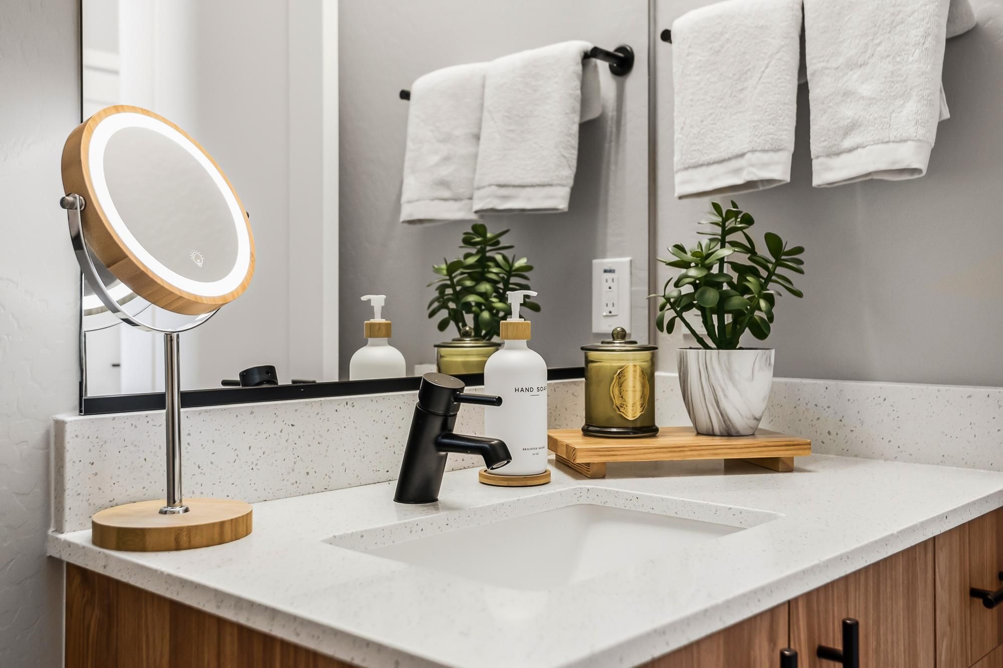Sonita apartments Modern bathroom vanity with white countertop, plants, hand soap, towels, and a round lighted mirror.