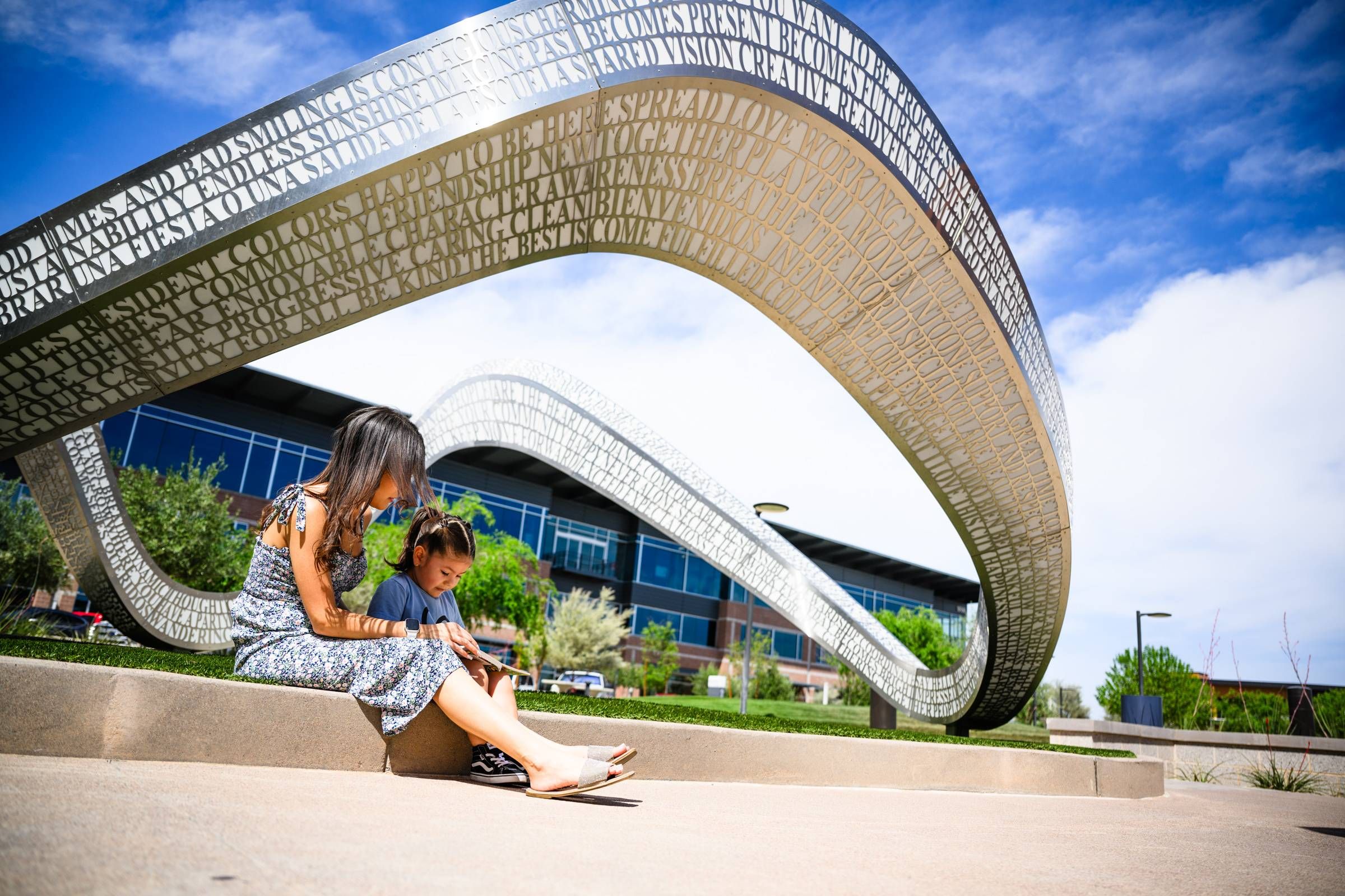 Sonita A woman and child sit reading on steps beneath a large, looping metallic sculpture with cut-out letters in Goodyear, AZ.