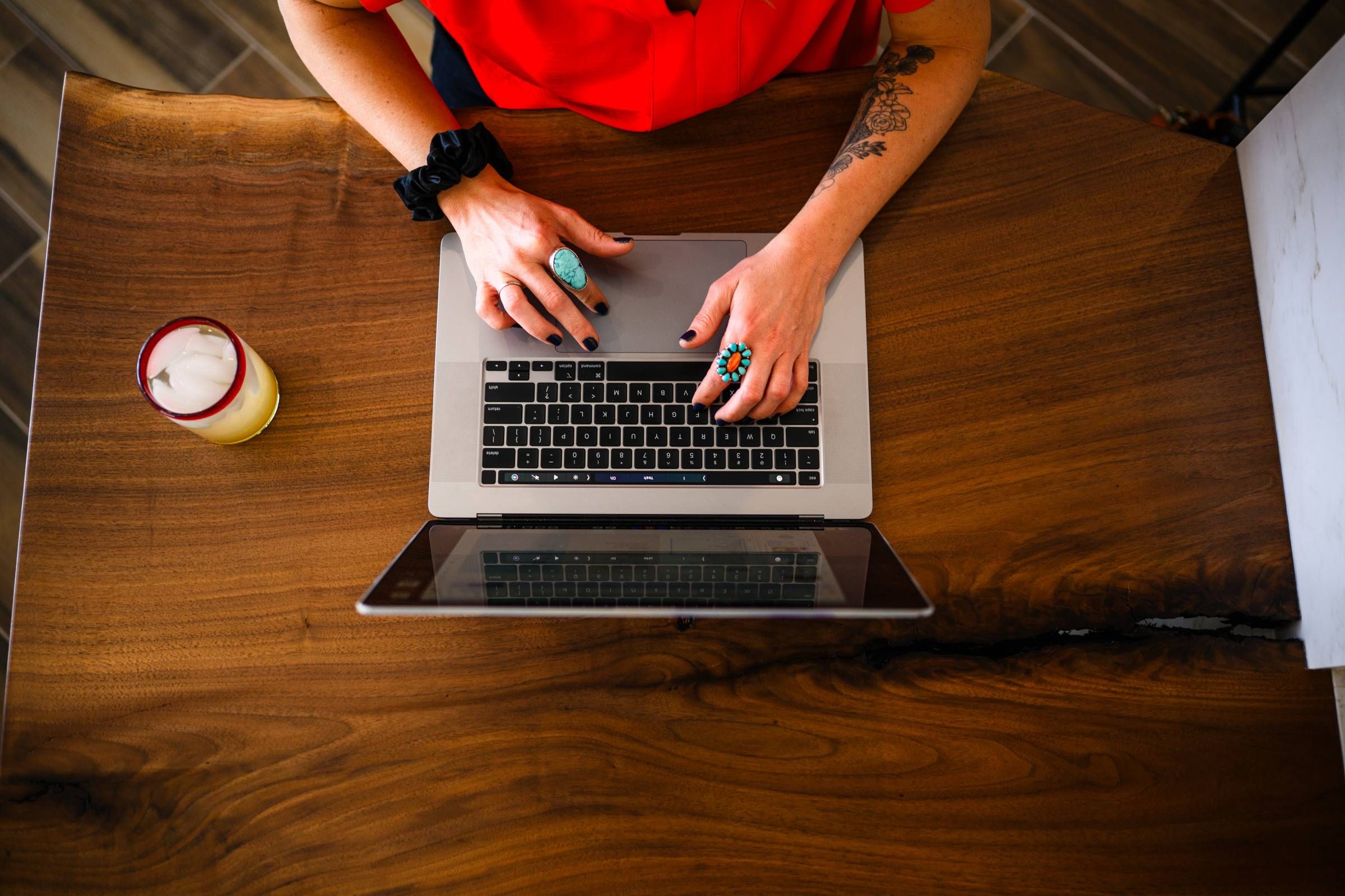 Sonita Person with rings types on a laptop at a wooden desk with a drink and ice beside them.