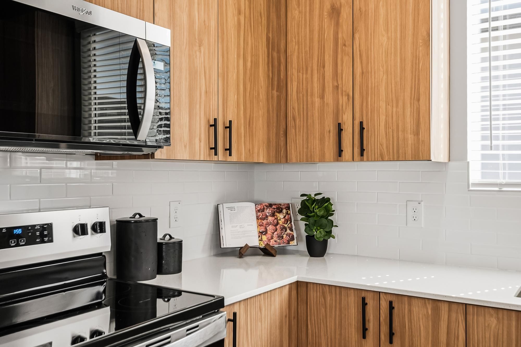 Sonita apartments Modern kitchen with wood cabinets, white counters, cookbook, plant, and stainless steel appliances.