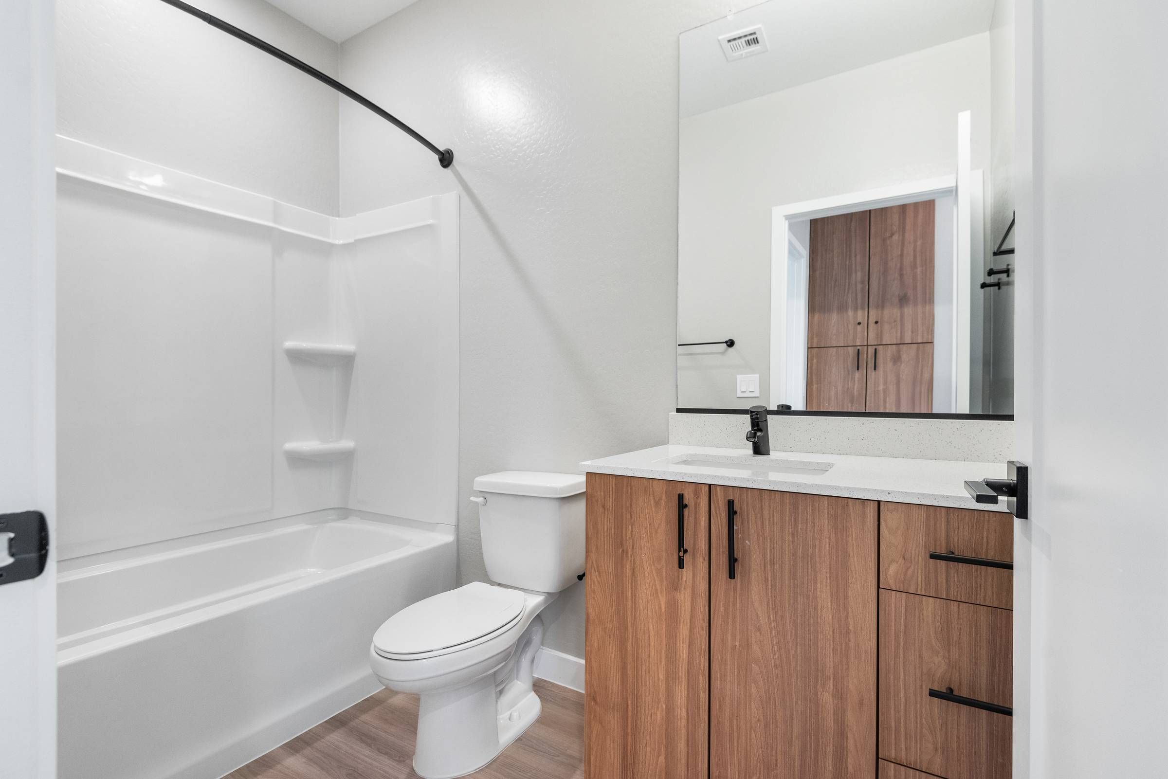 Sonita Apartments in Goodyear, AZ Modern bathroom with white bathtub, toilet, wood vanity, large mirror, and light gray walls.