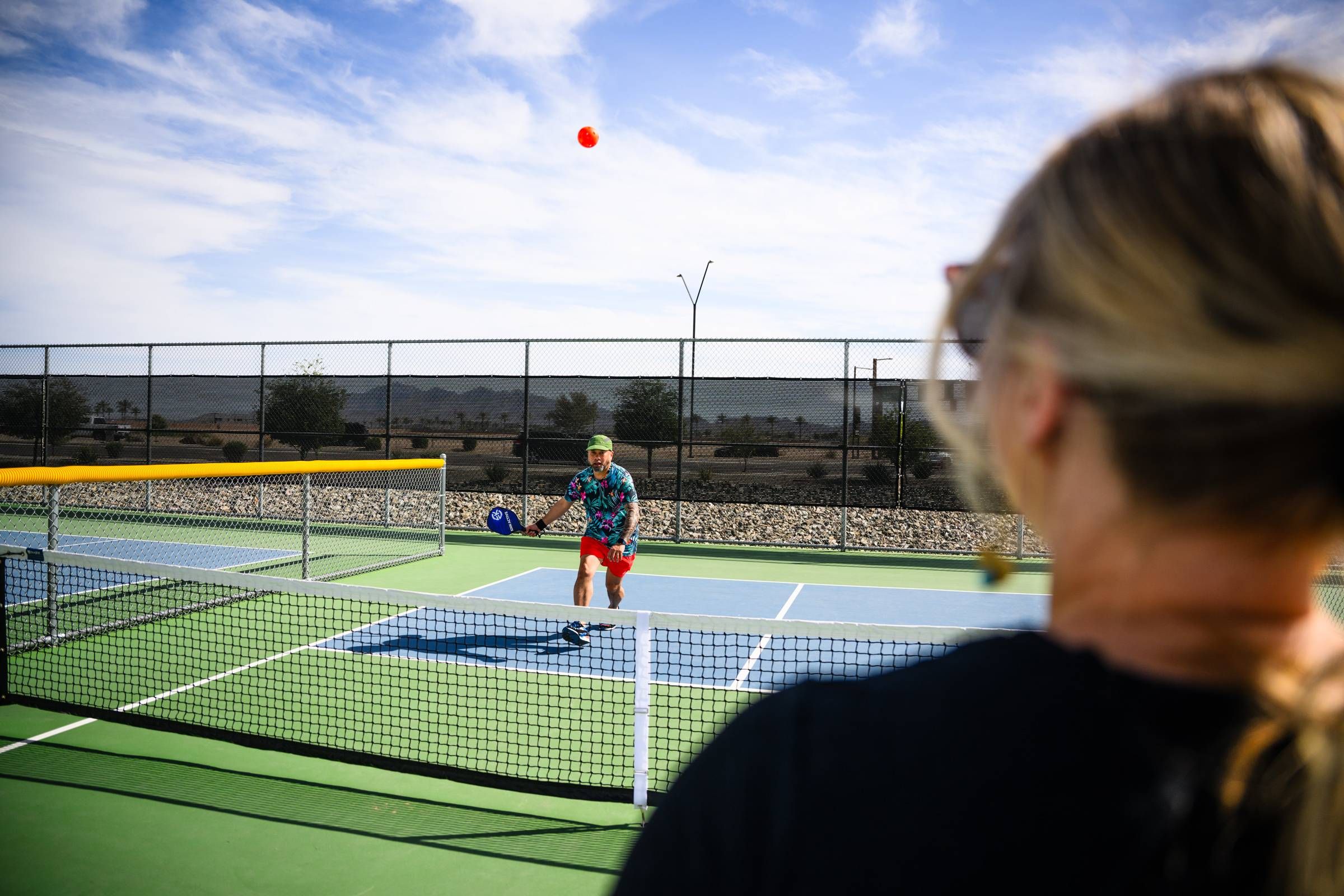 A person plays pickleball outdoors on a sunny day, as another watches from behind the net in Goodyear, AZ near Sonita Apartments.