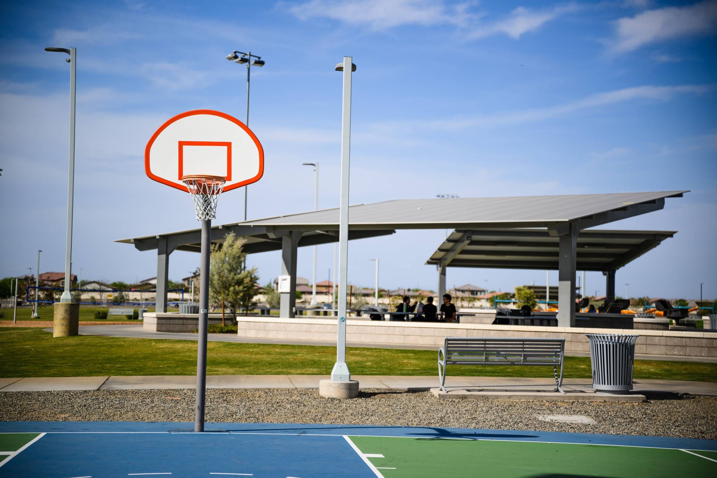 Outdoor basketball hoop on a court near a covered picnic area in a sunny park.