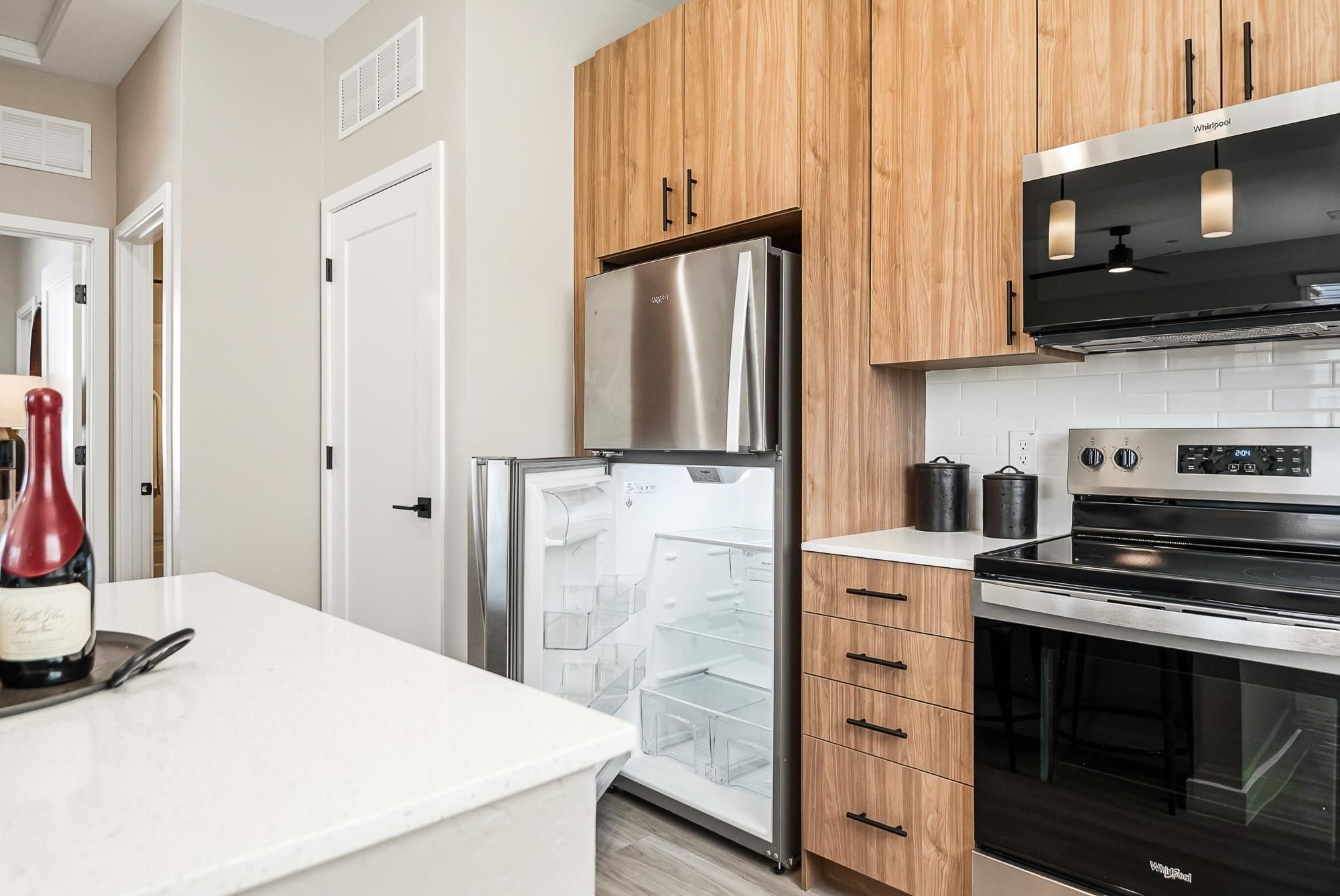 Sonita apartments Modern kitchen with wood cabinets, open stainless fridge, electric stove, and a bottle of wine on the counter.