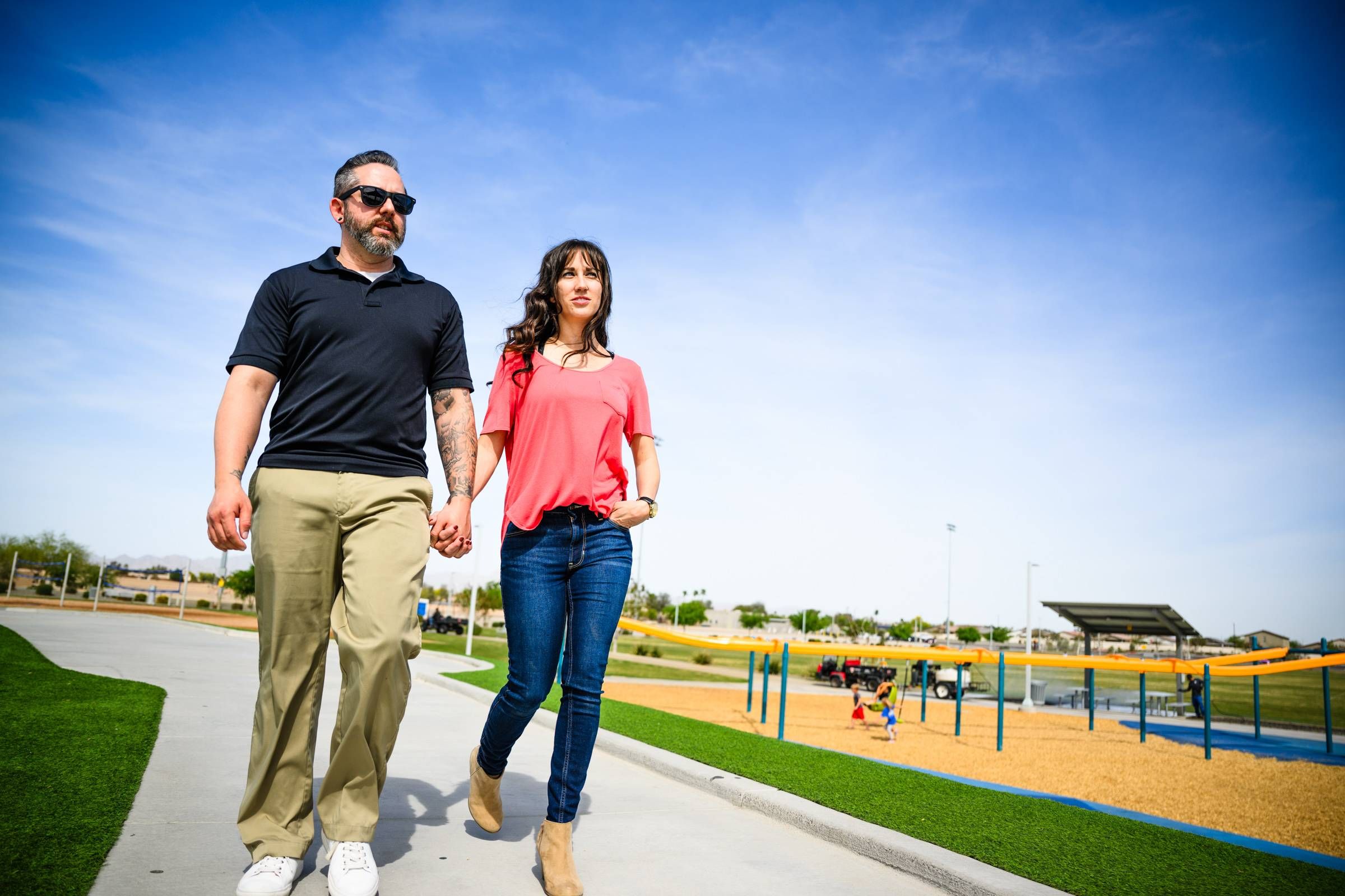 A man and woman hold hands while walking on a path in a sunny park with blue sky.