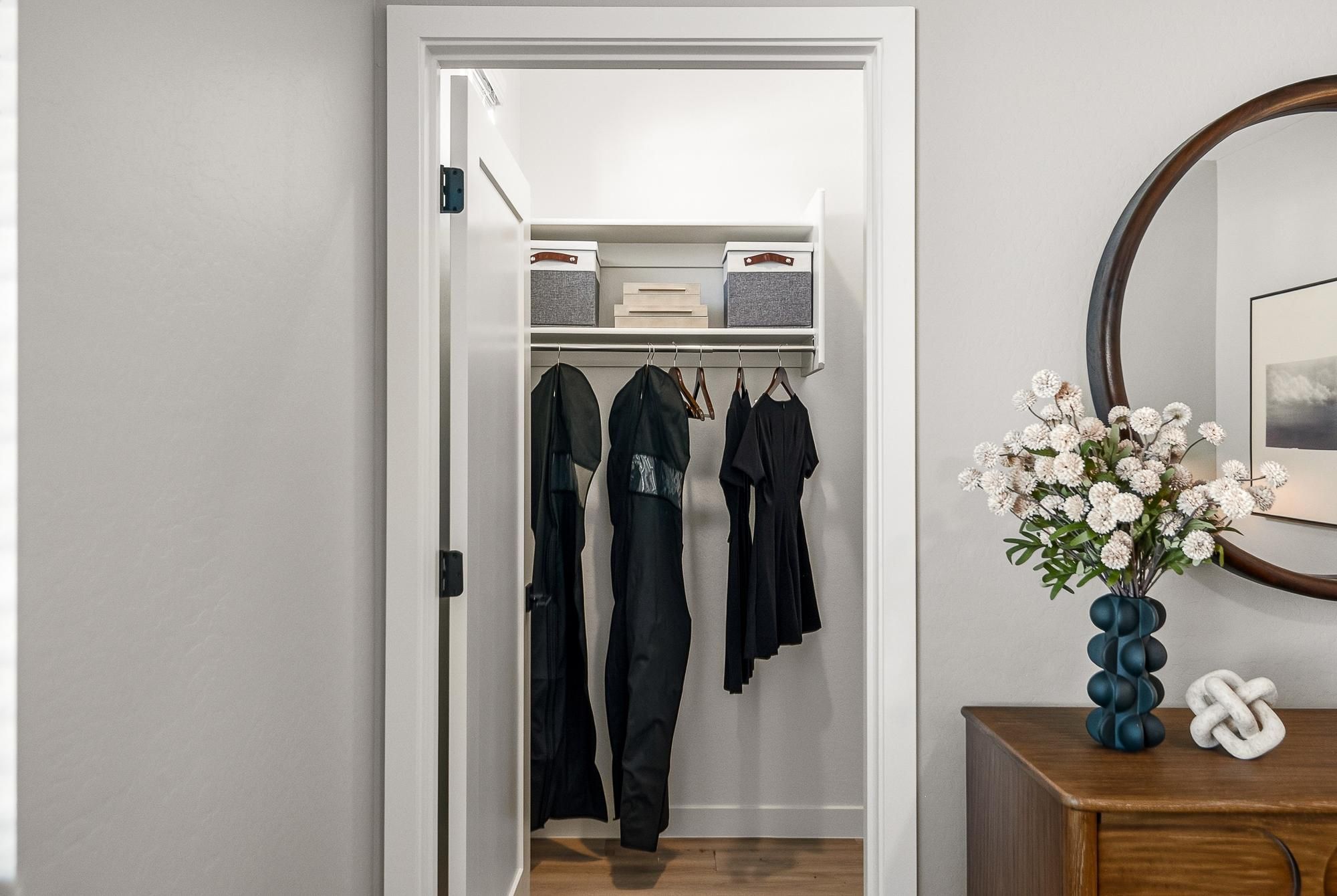 Sonita apartments Small closet with black clothes hanging, storage boxes above, next to a dresser with flowers and a round mirror.