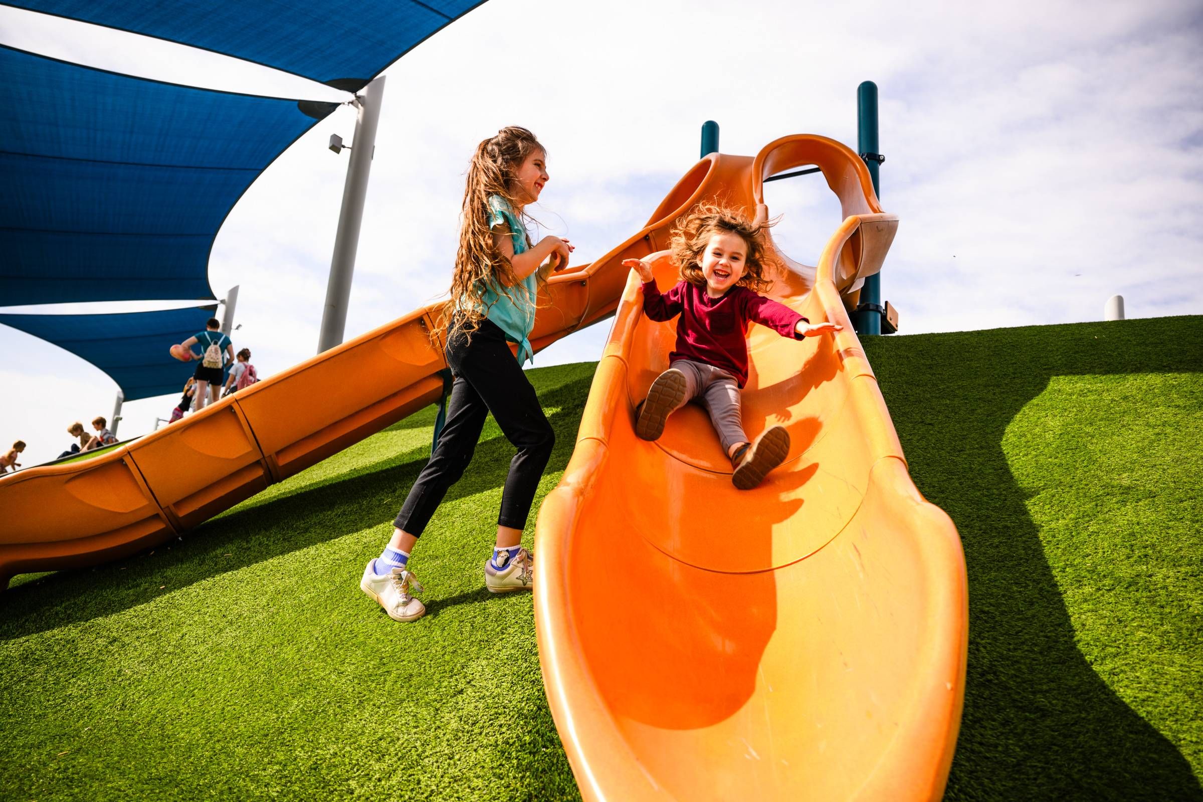 Two young girls play on an orange slide at a playground with green turf and blue shade canopies.