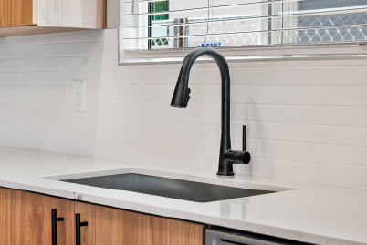 Sonita Modern kitchen sink with a black faucet, wood cabinets, and white tile backsplash under a window with blinds.